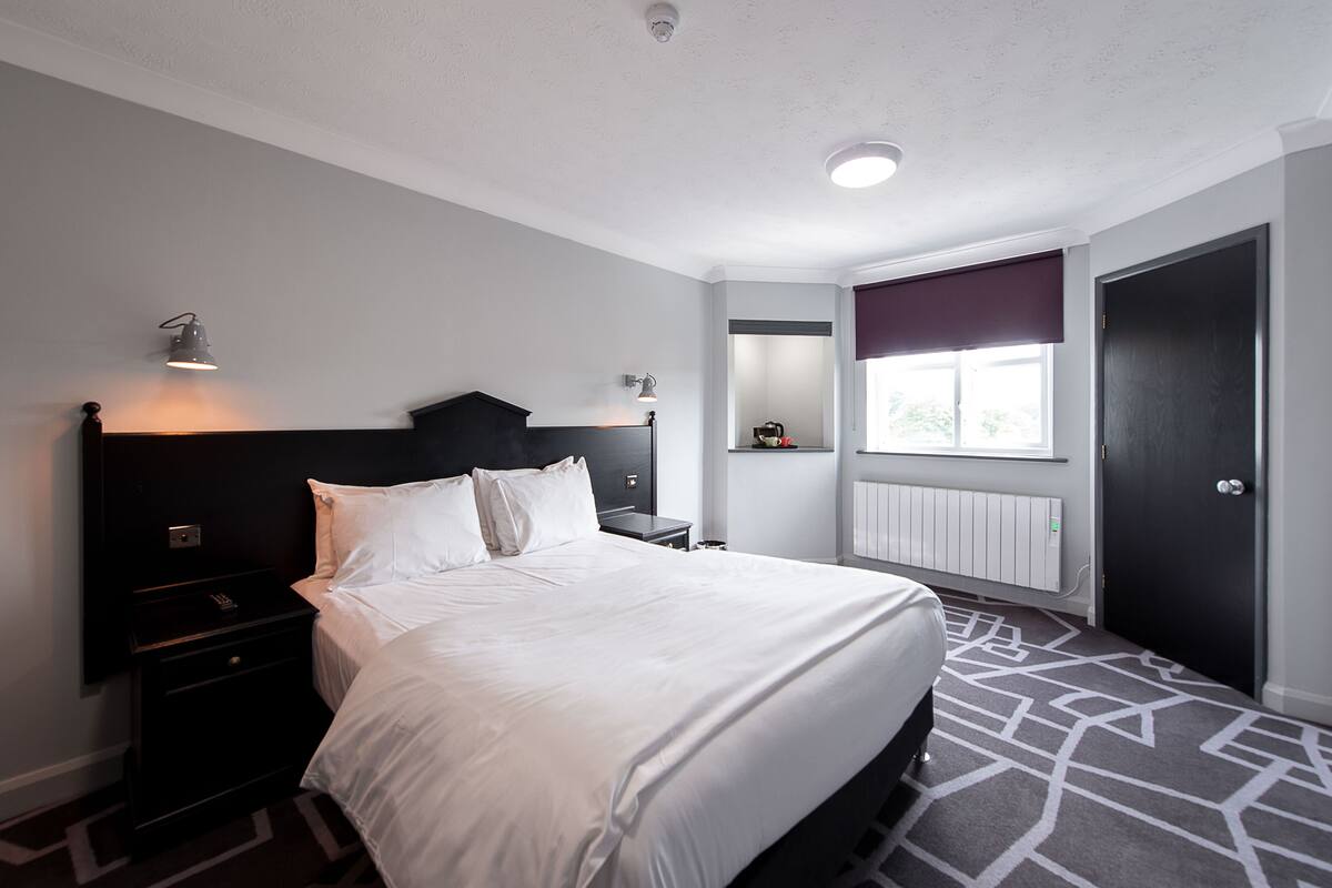 A modern hotel room with a large bed, bedside tables, a nightstand, a window with a view, and a door.