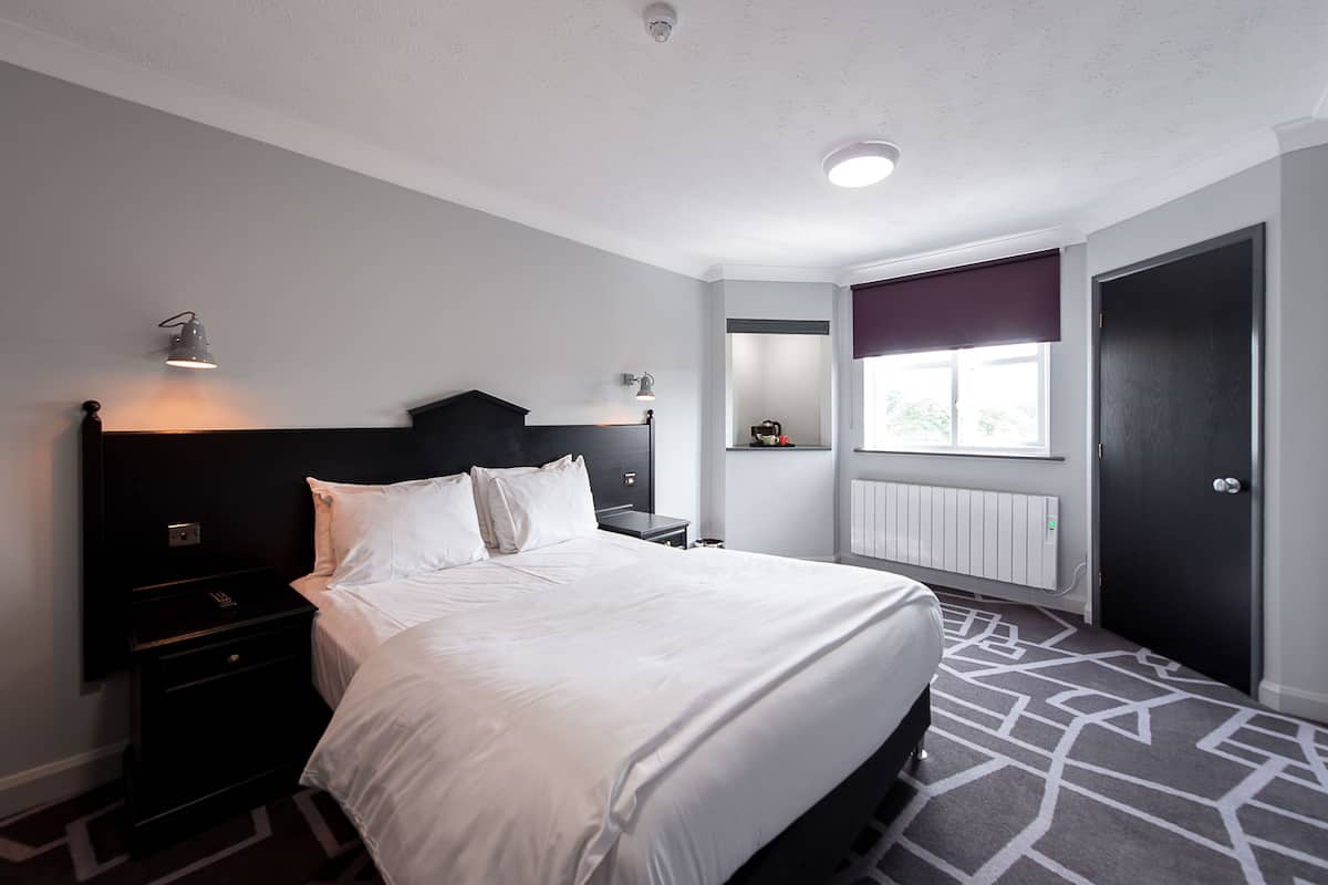 A modern hotel room with a large bed, bedside tables, a nightstand, a window with a view, and a door.