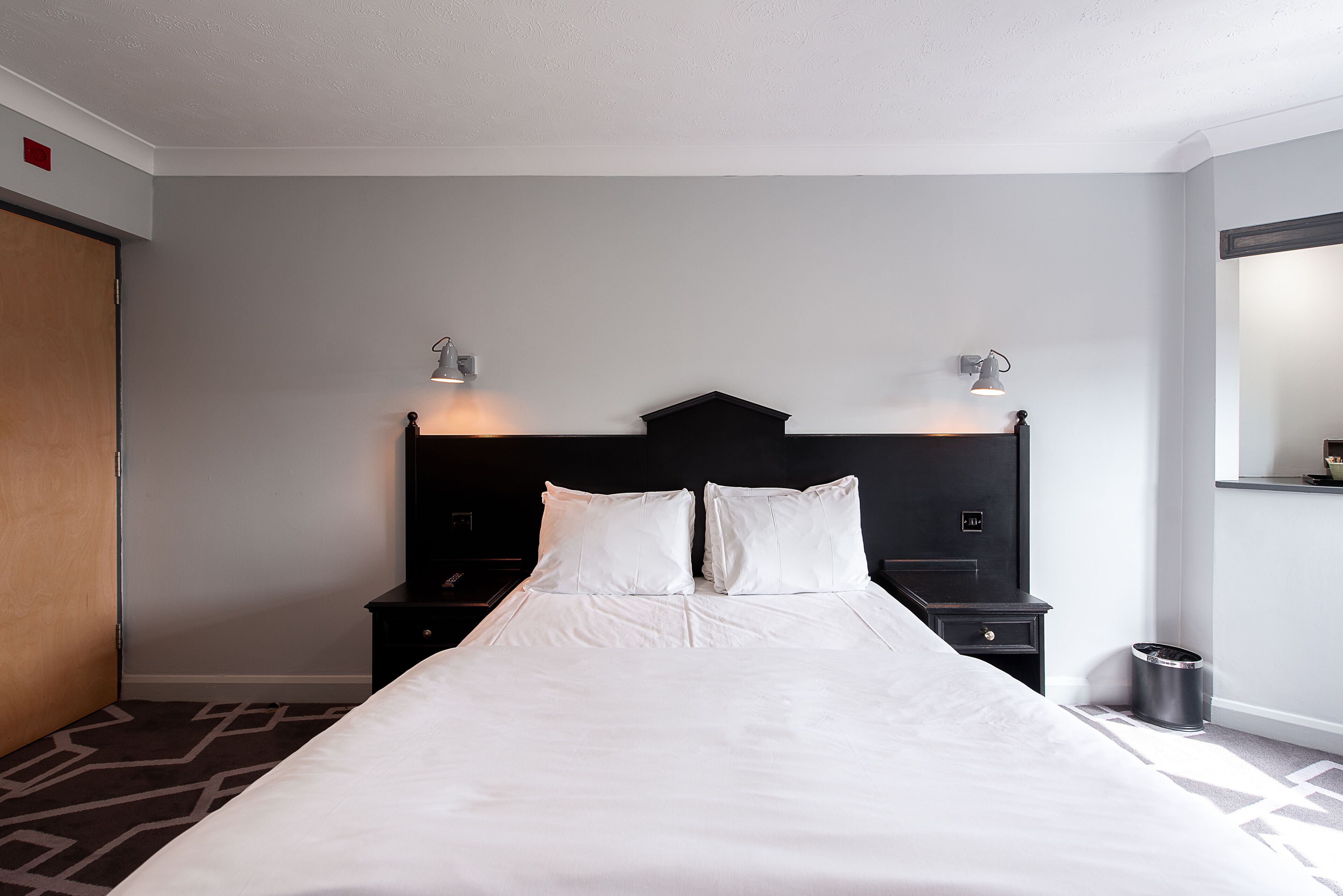 Habitación de hotel con una cama grande, dos mesitas de noche, cabecera negra y una lámpara de pared.