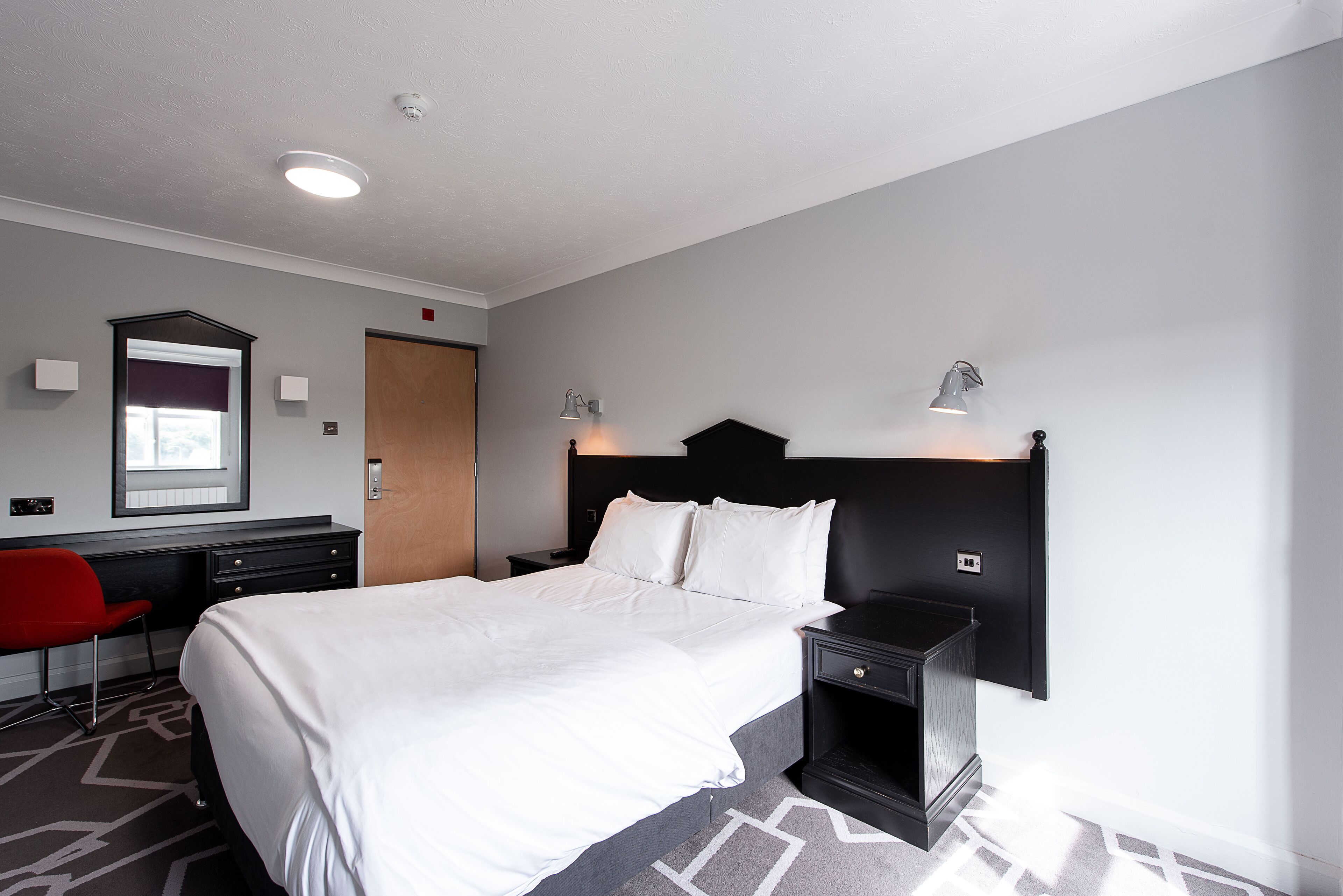 A hotel room with a large bed, a desk with a chair, a nightstand, and a wall-mounted lamp.