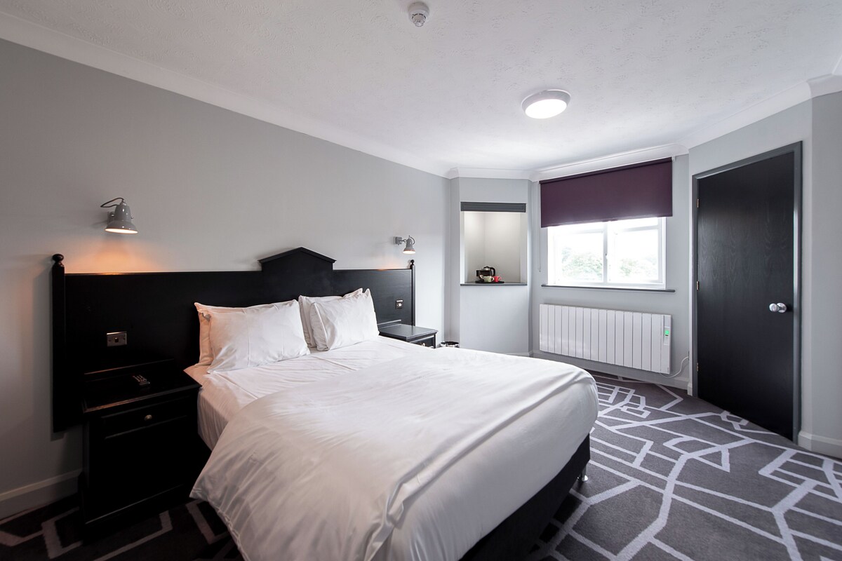 A modern hotel room with a large bed, bedside tables, a nightstand, a window with a view, and a door.