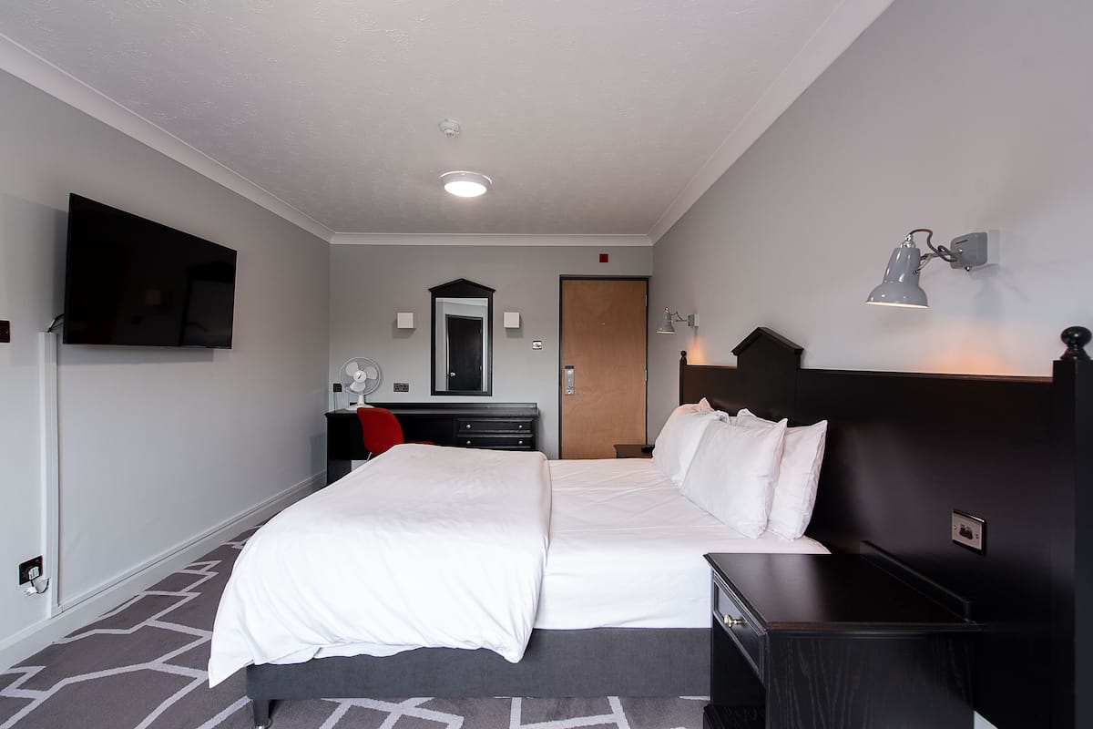 A hotel room with a large bed, a desk with a chair, a flat-screen TV, a mirror, and a wall-mounted lamp.