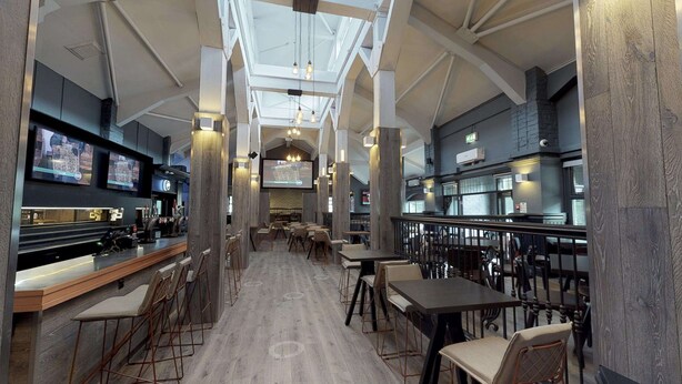 Un restaurant moderne avec un intérieur élégant, caractérisé par de hauts plafonds, des poutres apparentes et un bar central doté de plusieurs écrans.