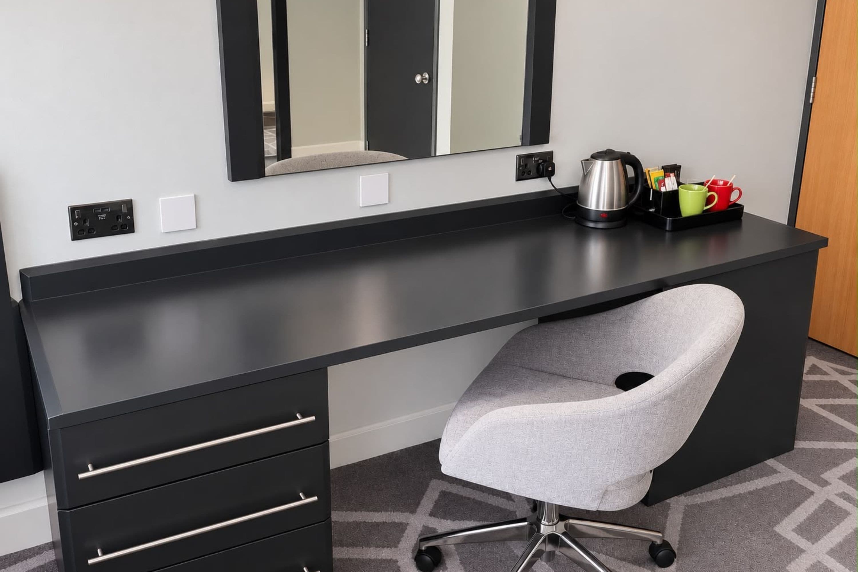 A modern hotel room with a black desk, a grey office chair, a large mirror, and a kettle on the desk.