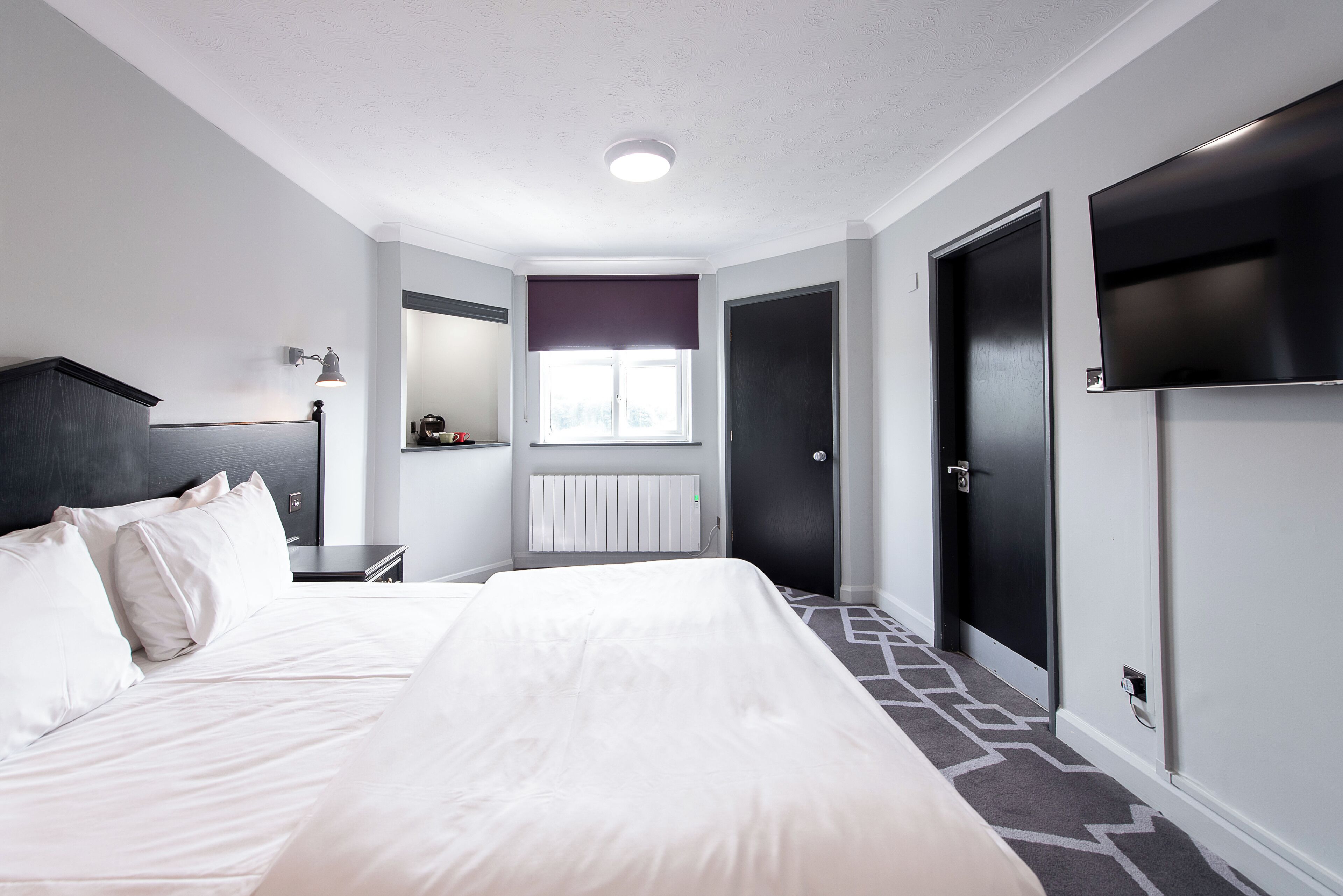 A hotel room with a large bed, a bedside table, a lamp, a window with blinds, a radiator, and a TV mounted on the wall.