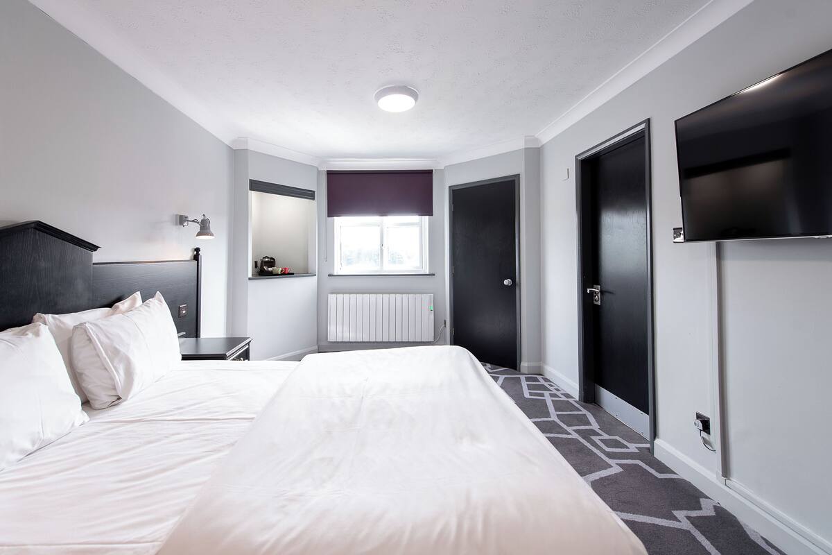 A hotel room with a large bed, a bedside table, a lamp, a window with blinds, a radiator, and a TV mounted on the wall.