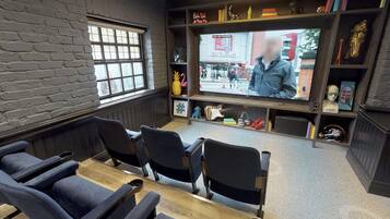 Habitación con una televisión mostrando una transmisión en vivo, una pared de ladrillo, una ventana y una estantería con diversos objetos.