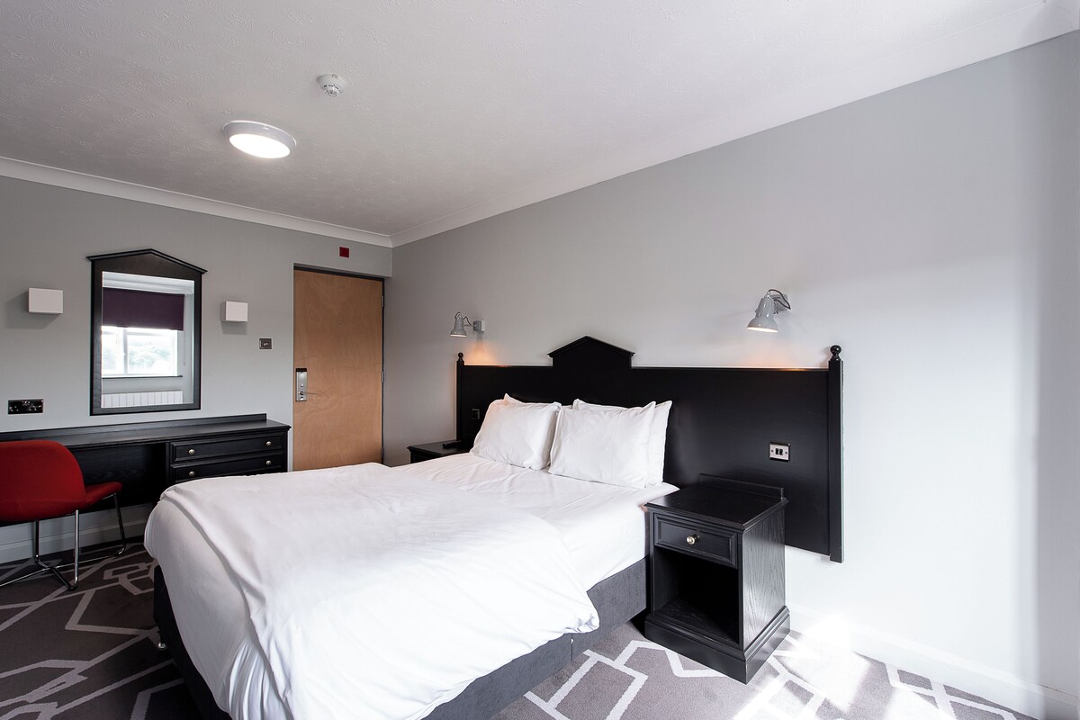 A hotel room with a large bed, a desk with a chair, a nightstand, and a wall-mounted lamp.