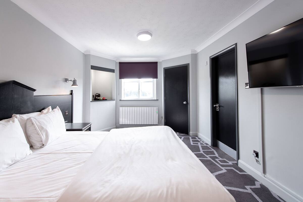 A hotel room with a large bed, a bedside table, a lamp, a window with blinds, a radiator, and a TV mounted on the wall.