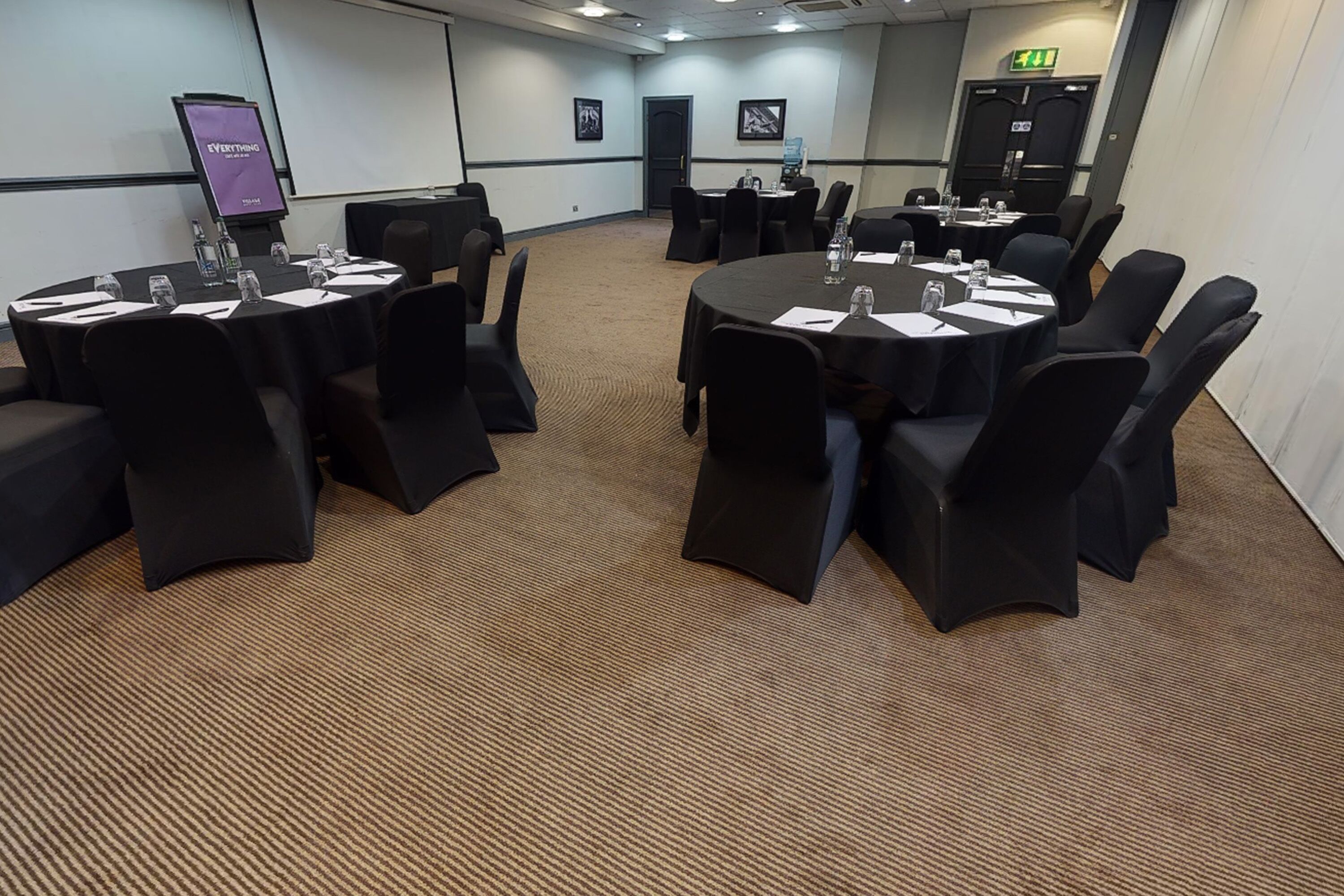 A conference room with round tables, black chairs, and a projector screen.