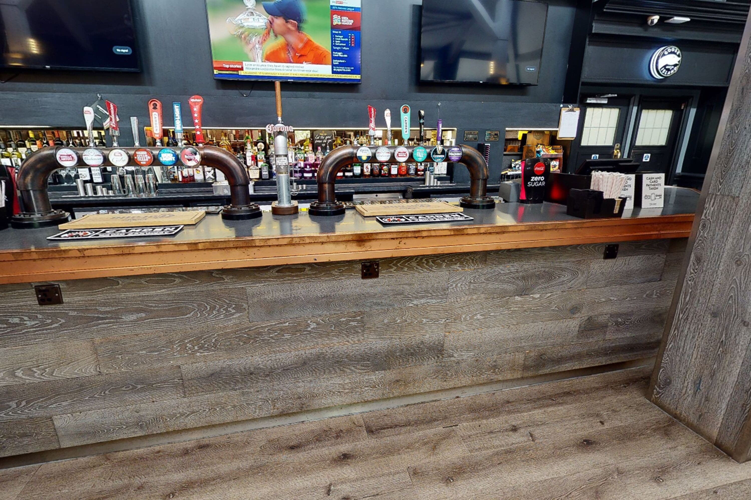 Zona de bar con mostrador de madera, varias grifos de cerveza y una pantalla de televisión arriba.