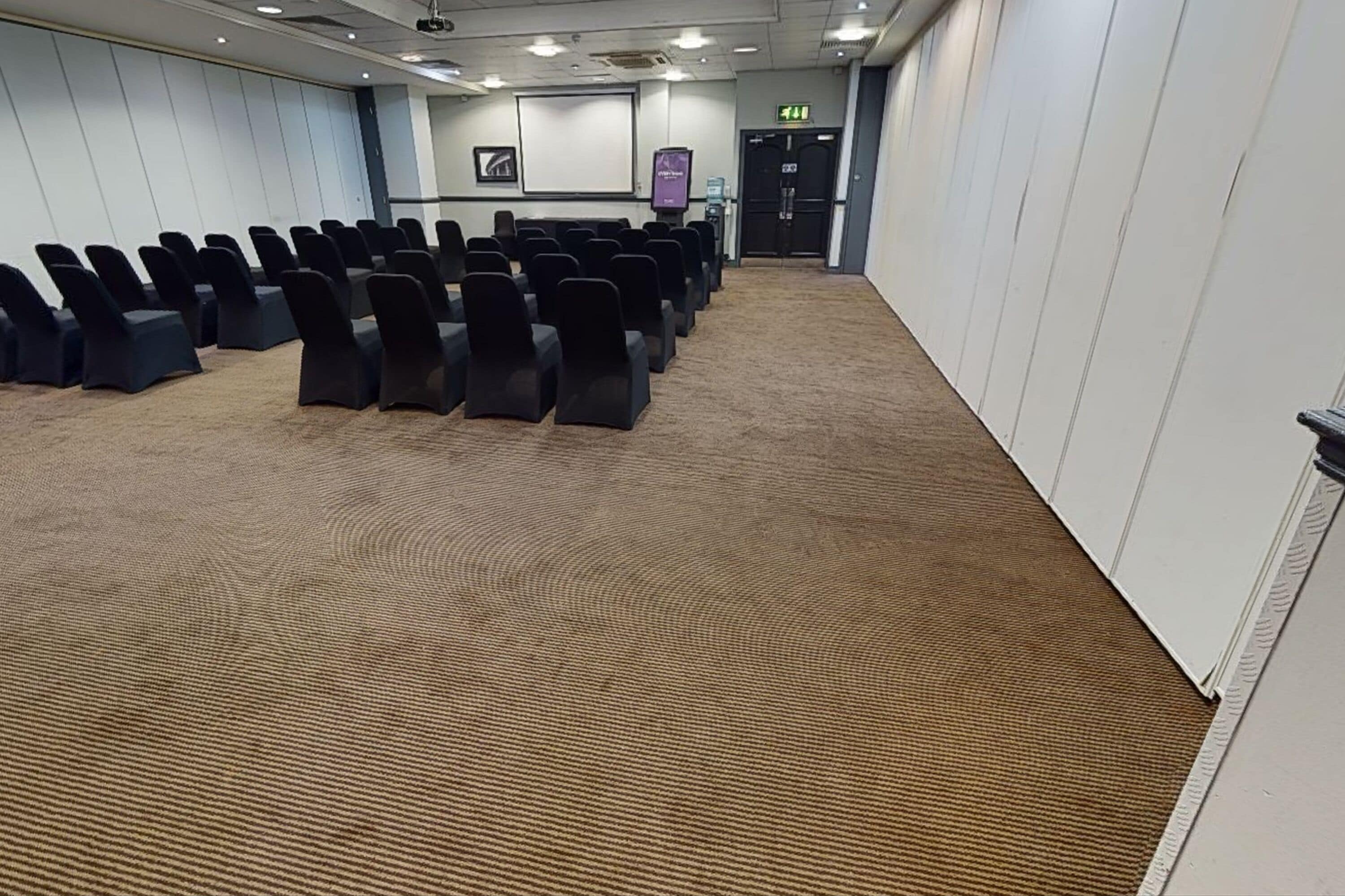 A conference room with rows of chairs, a carpeted floor, a projector screen, and a door with an exit sign.