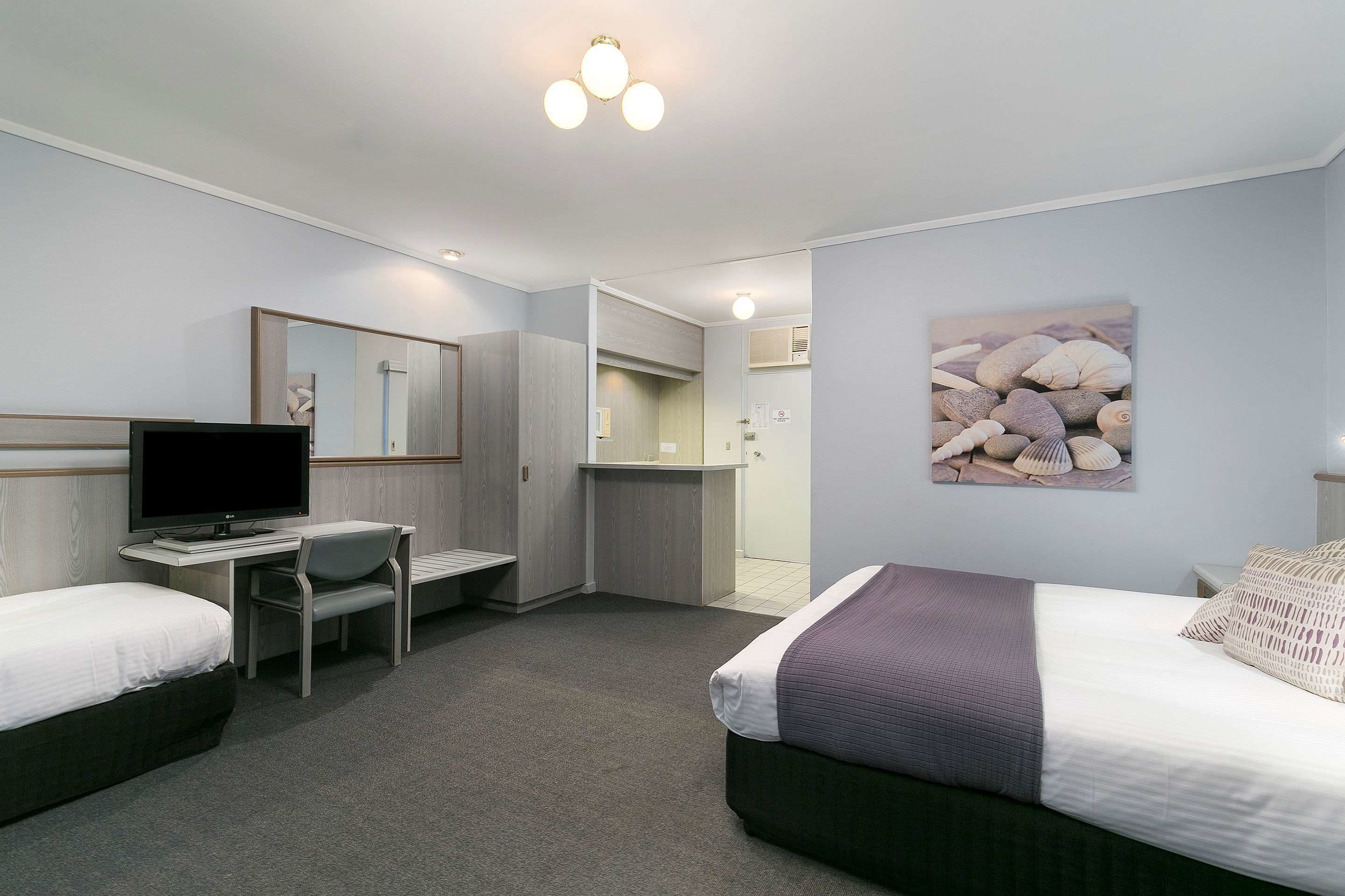 A hotel room with two beds, a desk with a computer, a mirror, and a painting of shells on the wall.