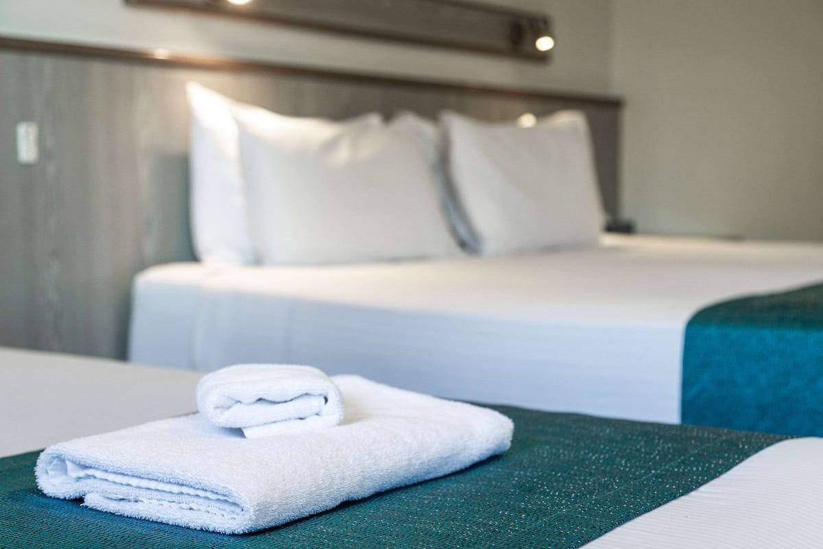 A neatly made bed with white linens and pillows, a folded white towel on a green surface, and a teal-colored bedspread.