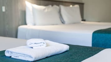 Una cama bien hecha con sábanas y almohadas blancas, una toalla blanca doblada sobre una superficie verde, y una colcha color aguamarina.