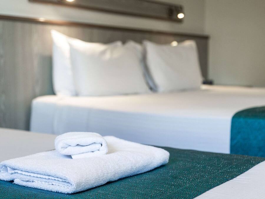 Een net opgemaakt bed met witte lakens en kussens, een gevouwen witte handdoek op een groen oppervlak, en een blauwgroene spreidekker.