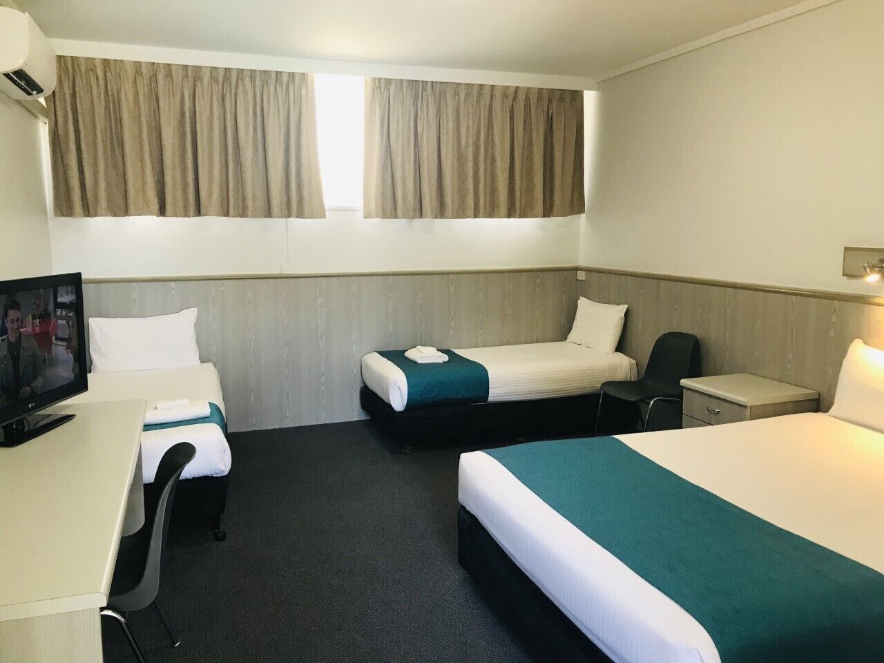 A hotel room with two single beds, a desk with a chair, a TV, and a window with curtains.