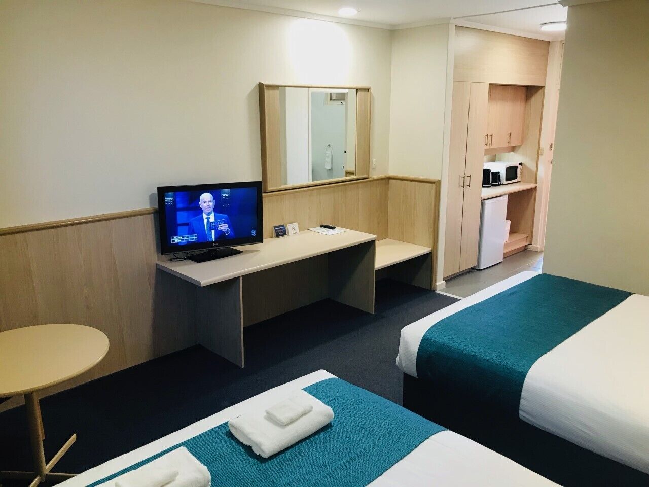 A hotel room with two beds, a desk with a computer, a mirror, a small table, and a kitchenette.