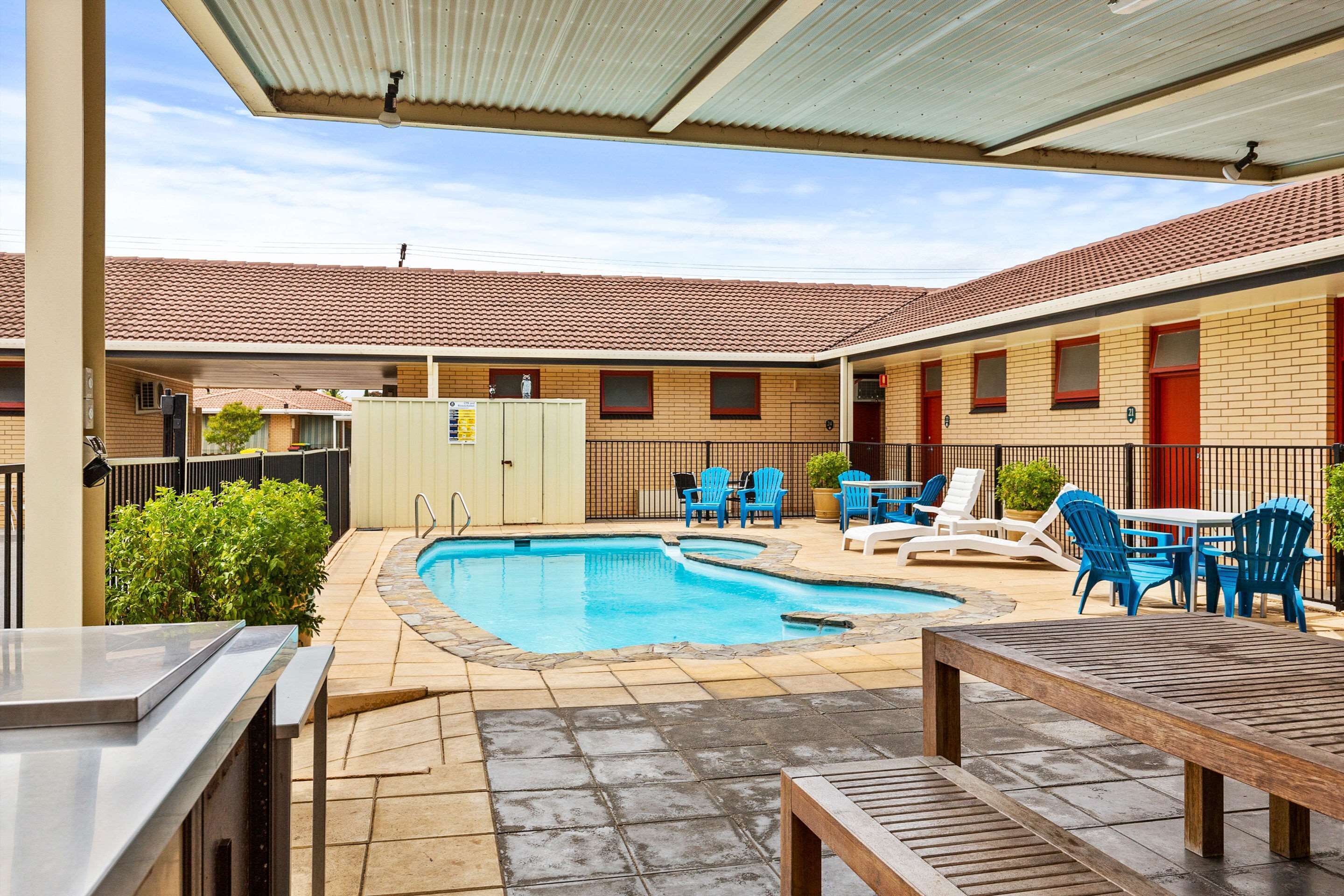 A pool area with blue chairs and a wooden table, surrounded by a building with a tiled roof and brick walls.
