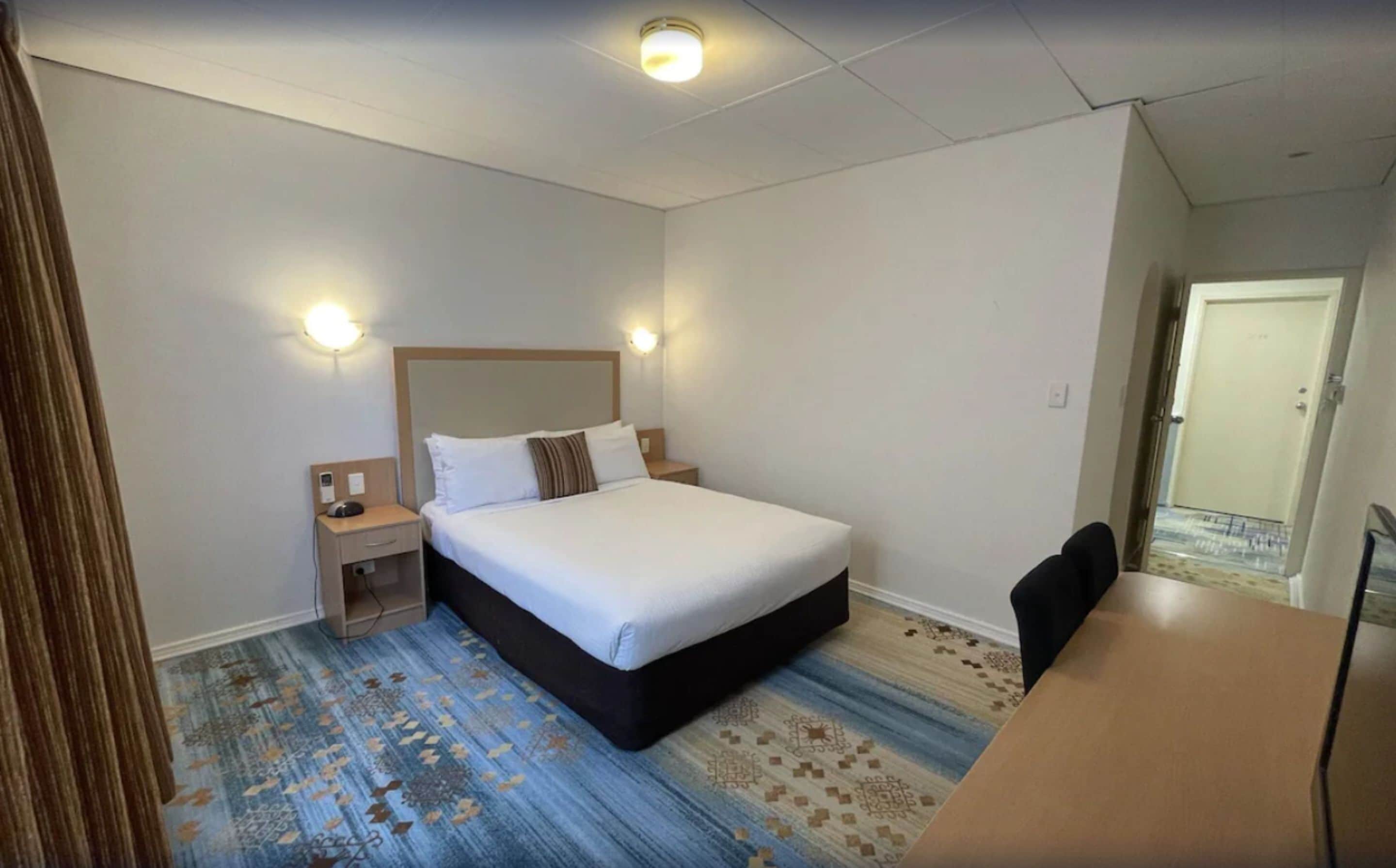 Et hotelværelse med en stor seng, sengeborde, et skrivebord og et mønstret tæppe.