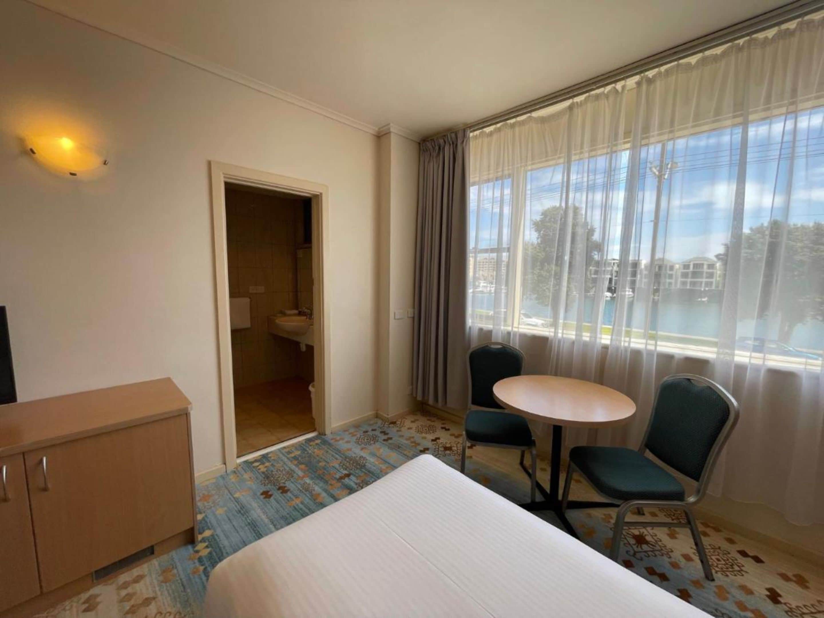 Habitación de hotel con cama, una pequeña mesa con dos sillas, un armario y un baño visible a través de una puerta abierta.