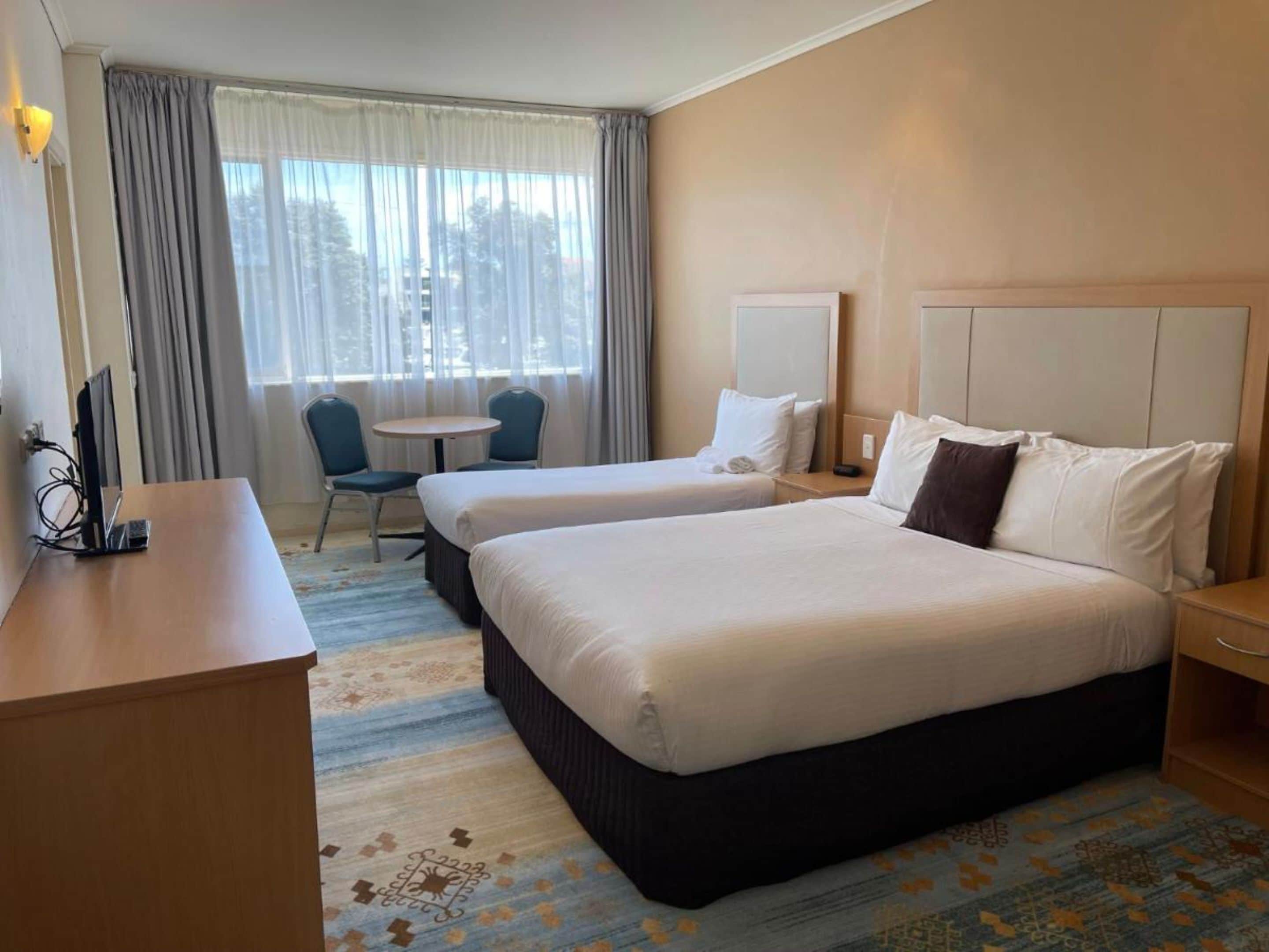 A hotel room with two beds, a desk, a chair, a small table, and a window with curtains.