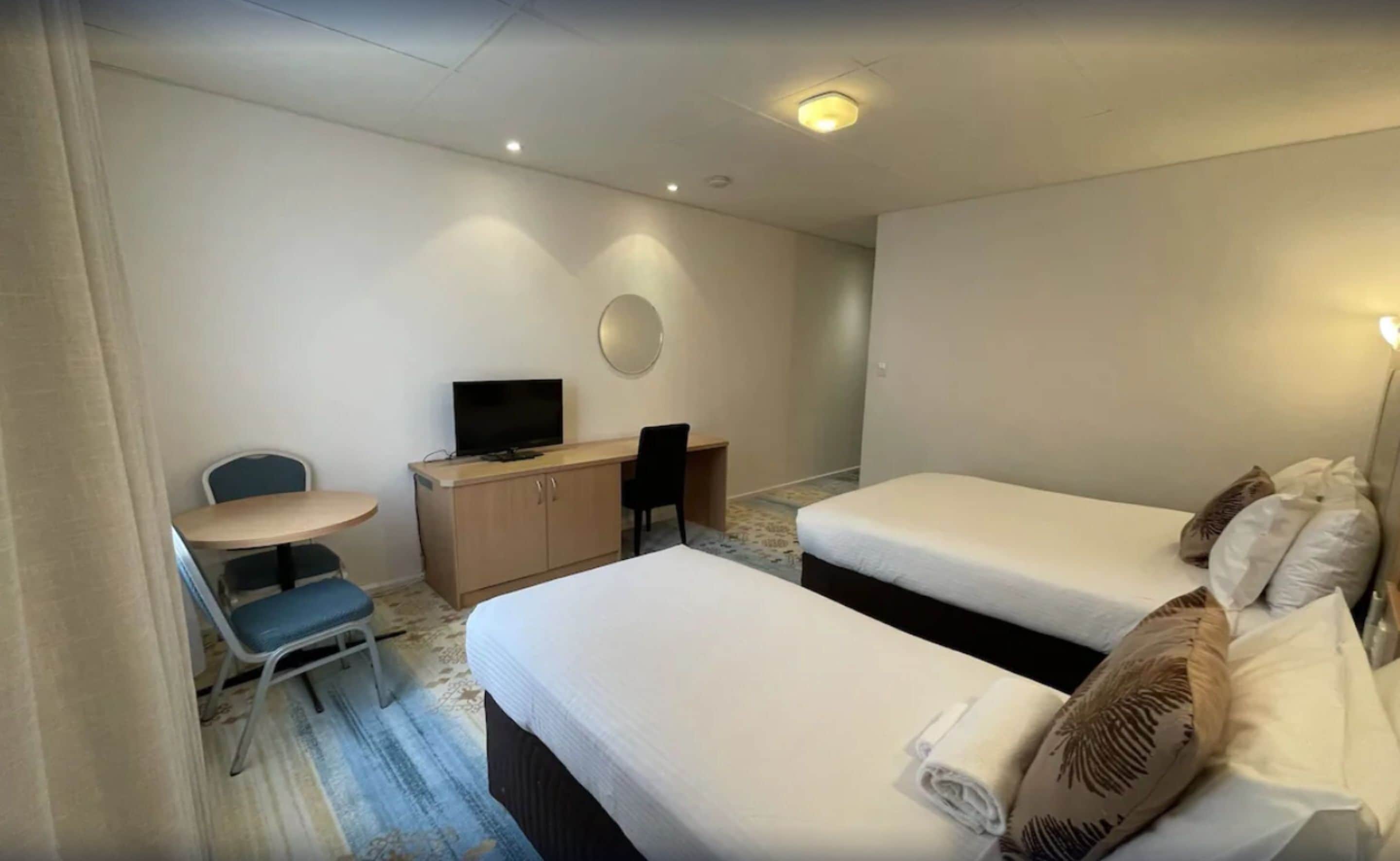 A hotel room with two single beds, a desk with a chair, a television, and a small table.