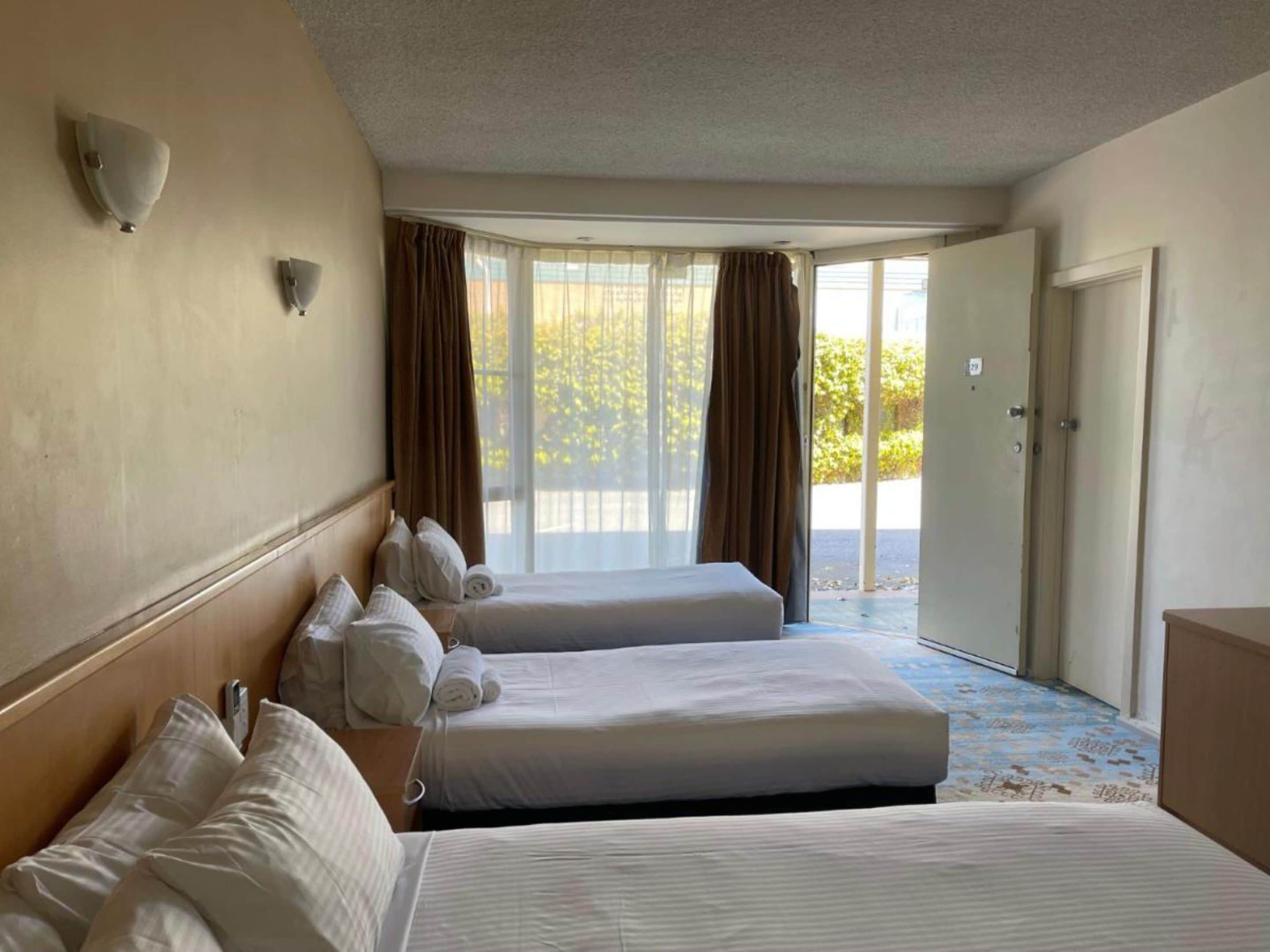 A hotel room with two beds, a wooden headboard, a large window with curtains, and a door leading outside.