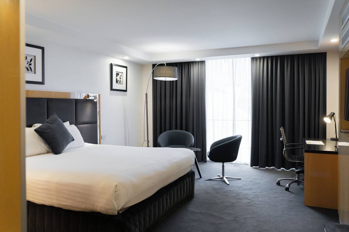 Et moderne hotelværelse med en stor seng, et skrivebord, en stol og et stort vindue med gardiner.