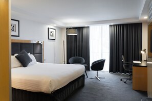 A modern hotel room with a large bed, a desk, a chair, and a large window with curtains.