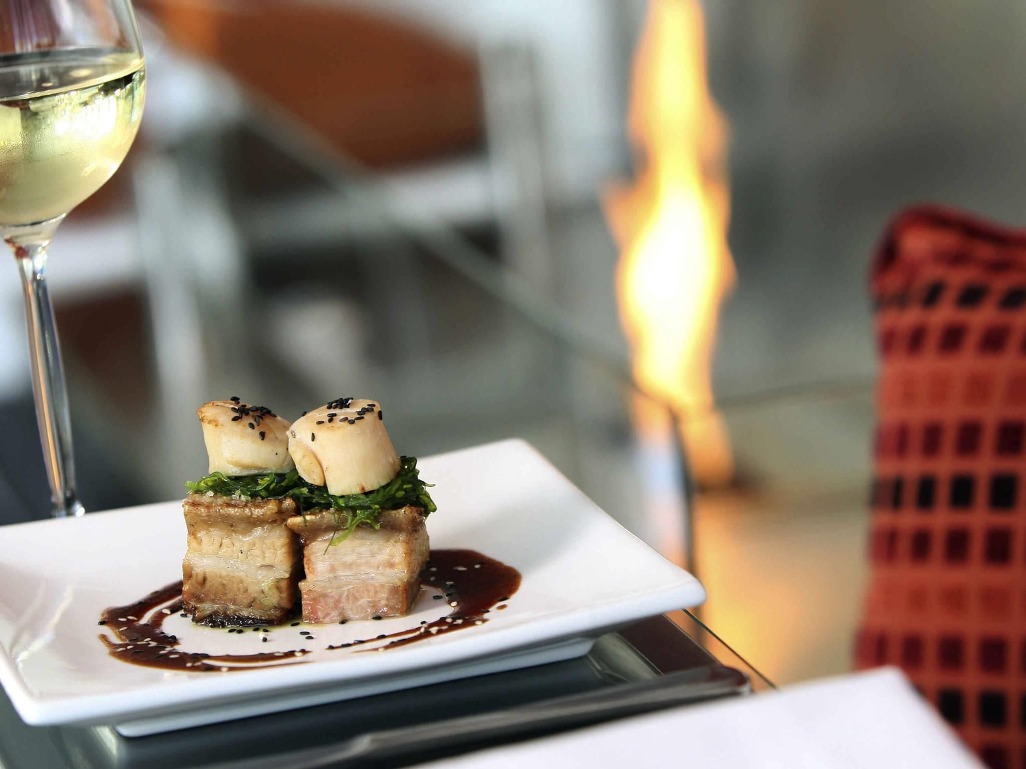 Un plato cuadrado con un guiso de vieiras a la plancha sobre una cama de hojas verdes, acompañado de una copa de vino blanco.