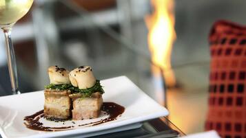 Un plato cuadrado con un guiso de vieiras a la plancha sobre una cama de hojas verdes, acompañado de una copa de vino blanco.