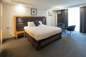 A hotel room with a large bed, a bedside table, a chair, and a large window with curtains.