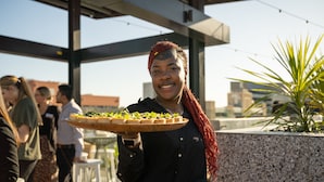 Una persona sostiene una bandeja con comida, ubicada en un espacio al aire libre con una estructura arquitectónica moderna y una planta en maceta.