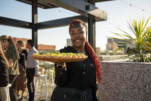 Una persona sostiene una bandeja con comida, ubicada en un espacio al aire libre con una estructura arquitectónica moderna y una planta en maceta.