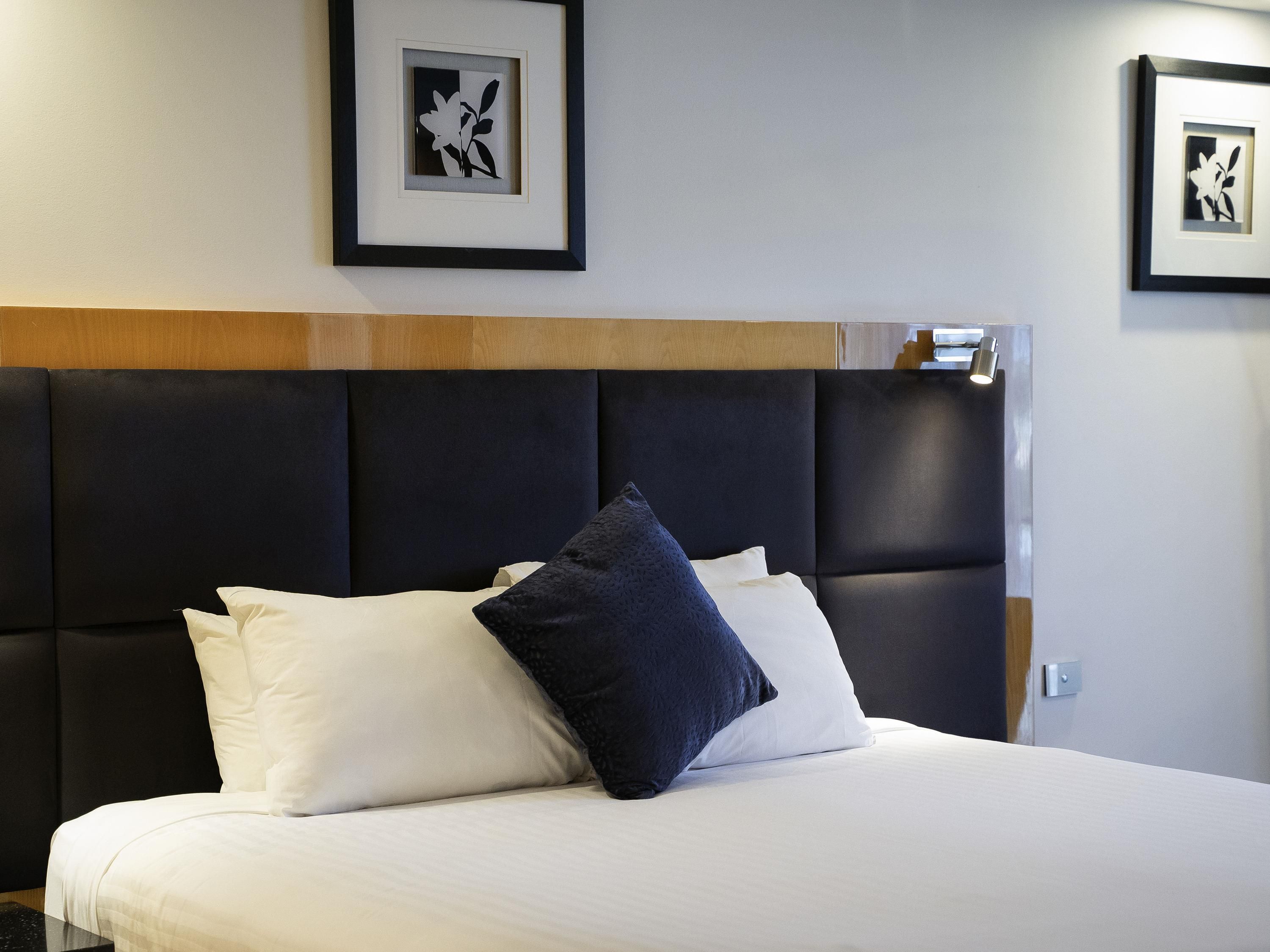 A neatly made bed with white and dark blue pillows, a dark headboard, and framed artwork on the wall.
