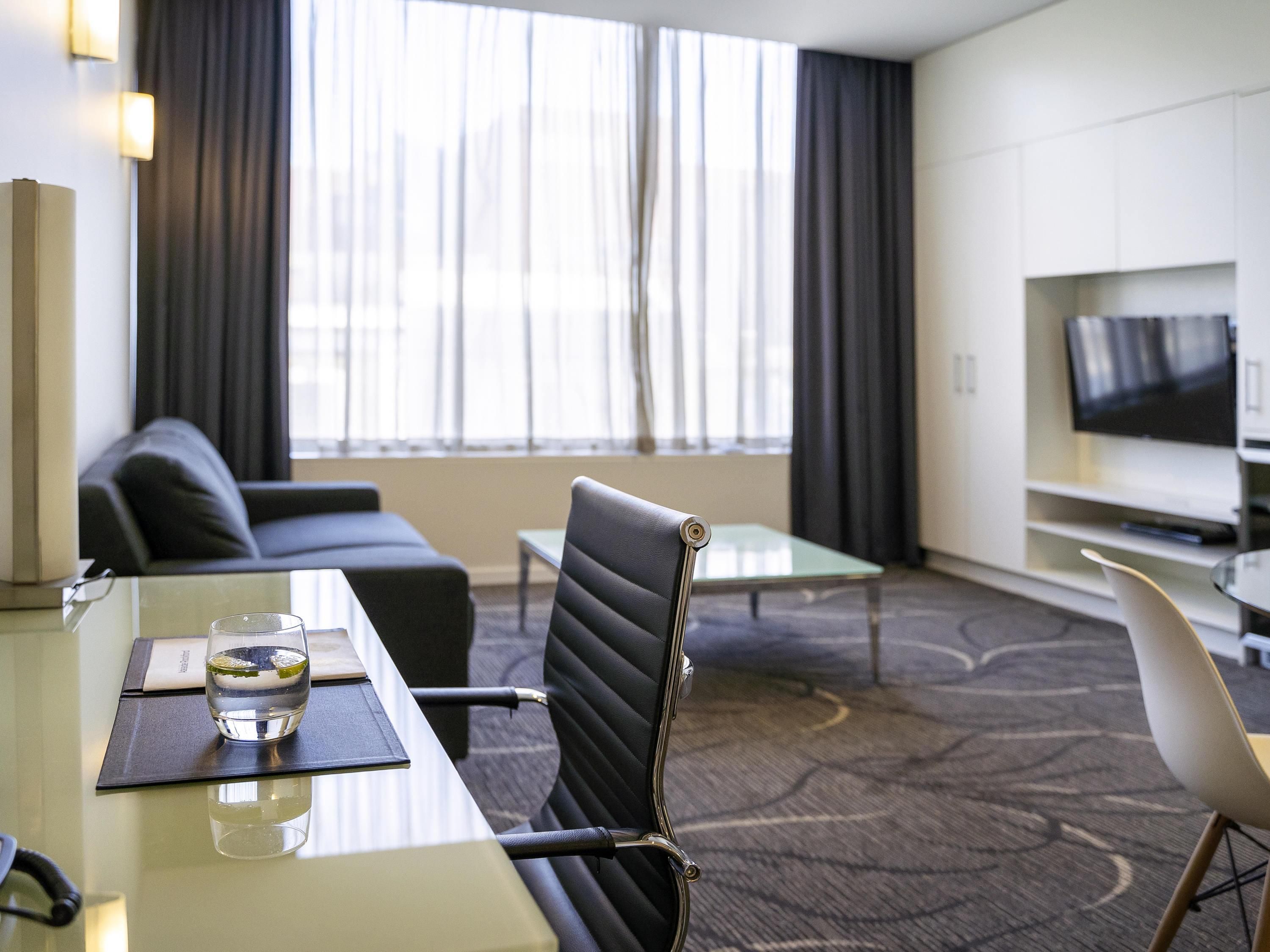Habitación de hotel con una mesa de comedor, sillas, un sofá, un televisor y un amplio ventanal con cortinas.