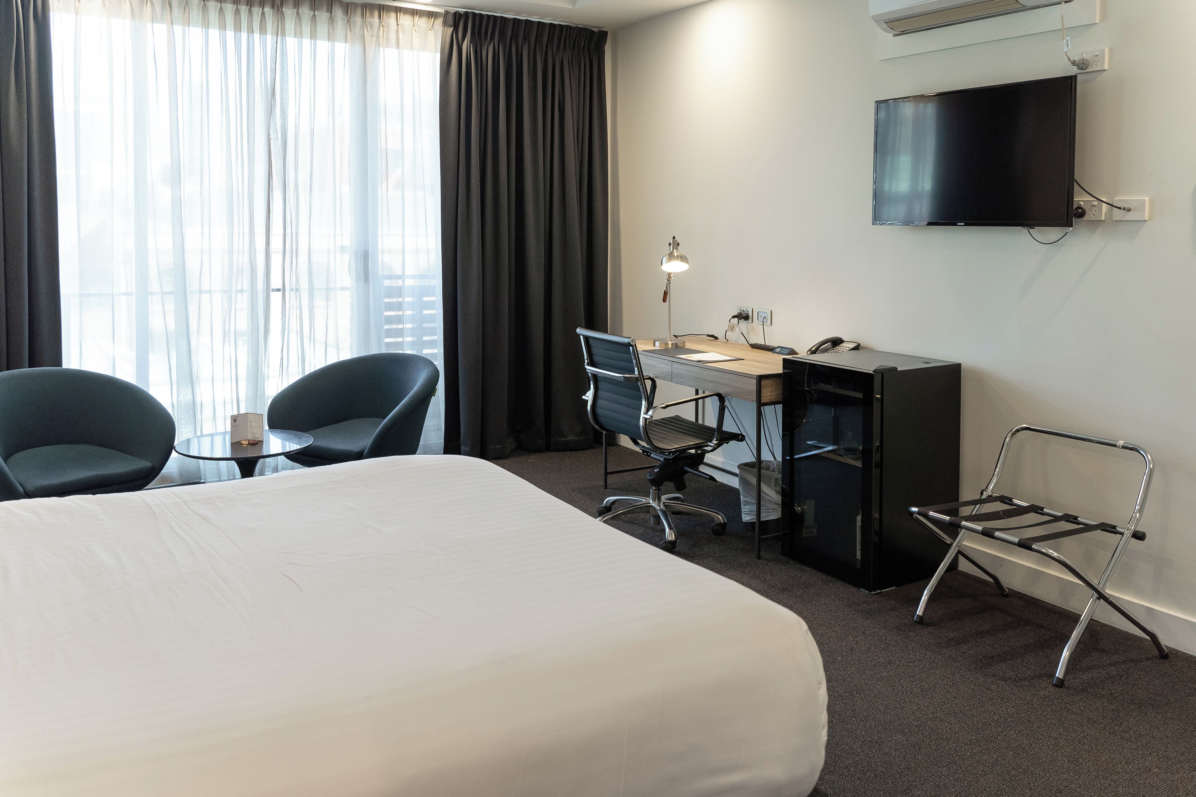 Une chambre d’hôtel avec un grand lit, deux chaises, un bureau, une télévision et une fenêtre avec des rideaux.
