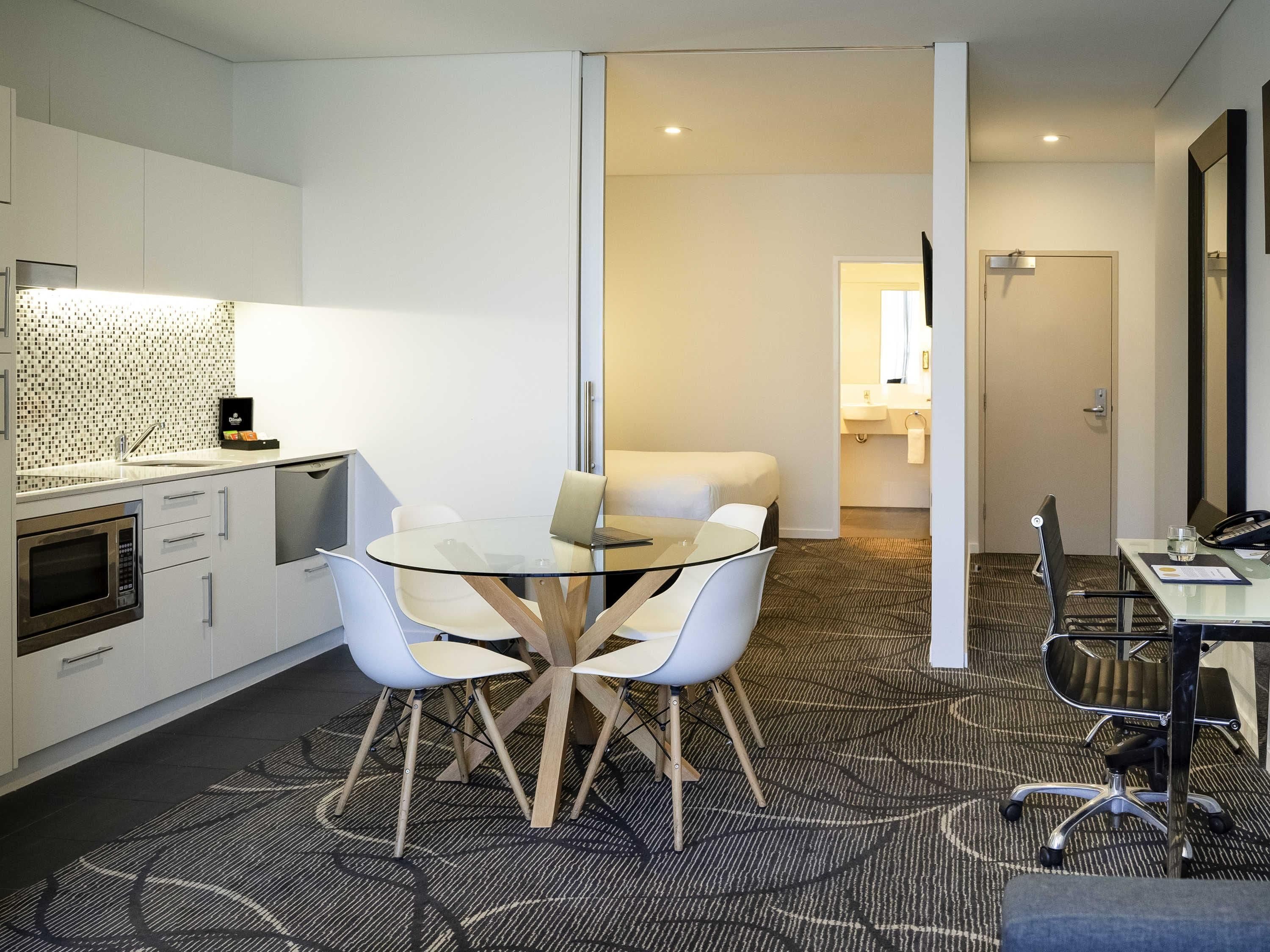 A modern hotel room with a glass dining table, white chairs, a kitchenette, a separate bathroom, and a carpeted floor.