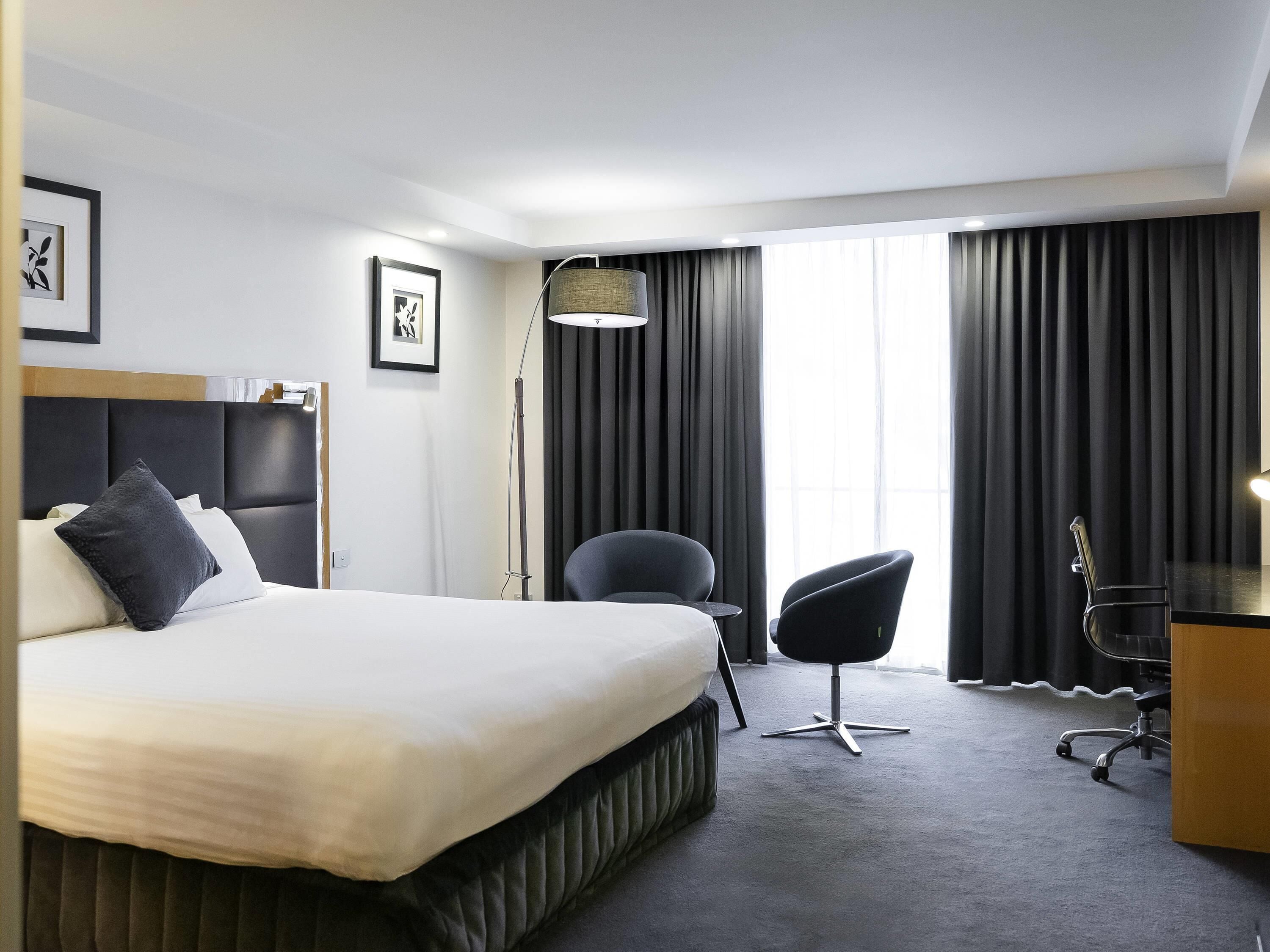 A hotel room with a large bed, two chairs, a desk, and a window with curtains.
