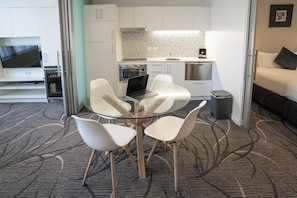 A modern hotel room with a glass dining table, white chairs, a laptop, a bed, a TV, and a kitchenette.