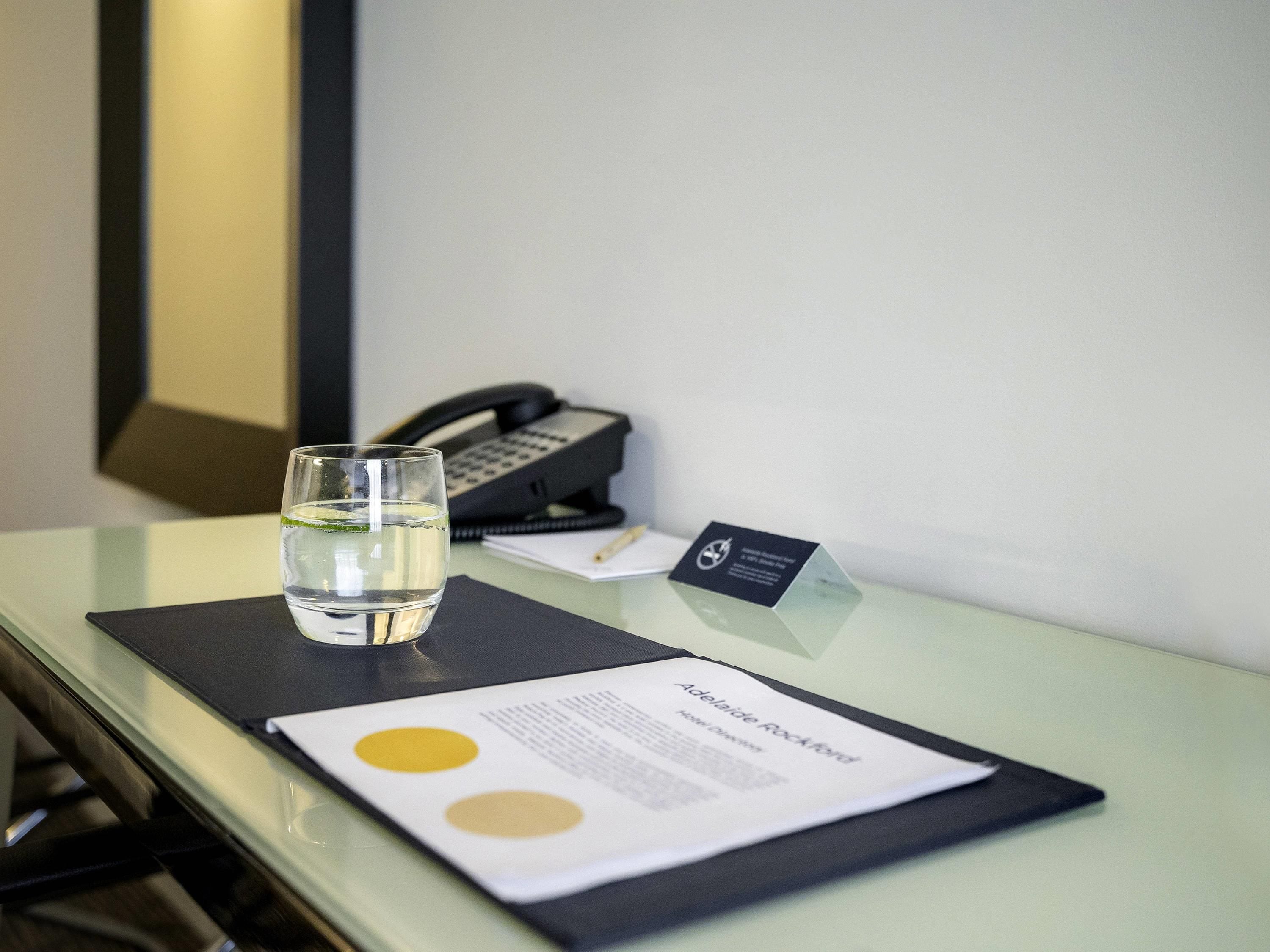 A hotel room with a glass of water, a telephone, a business card, and a menu on a table.