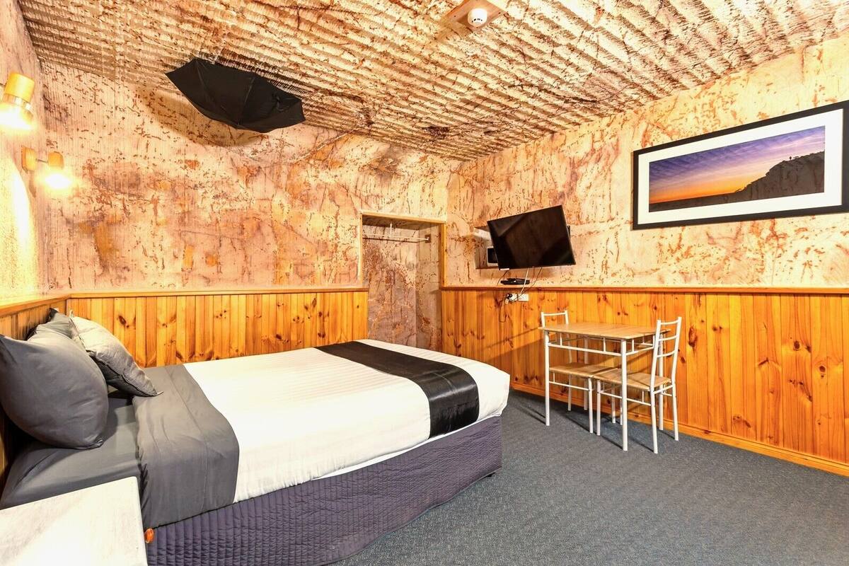A room with a bed, a desk, and a chair. There is a television on the wall and a framed picture above the desk.