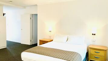 Quarto de hotel com cama, duas mesas de cabeceira, um criado-mudo, uma porta e piso coberto por carpete.