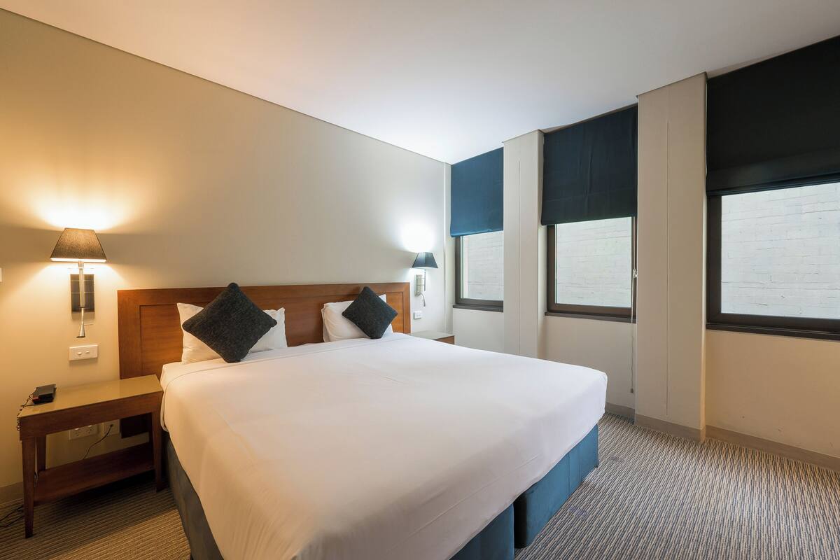 Quarto de hotel com uma cama grande, duas mesas de cabeceira e uma janela com persianas.