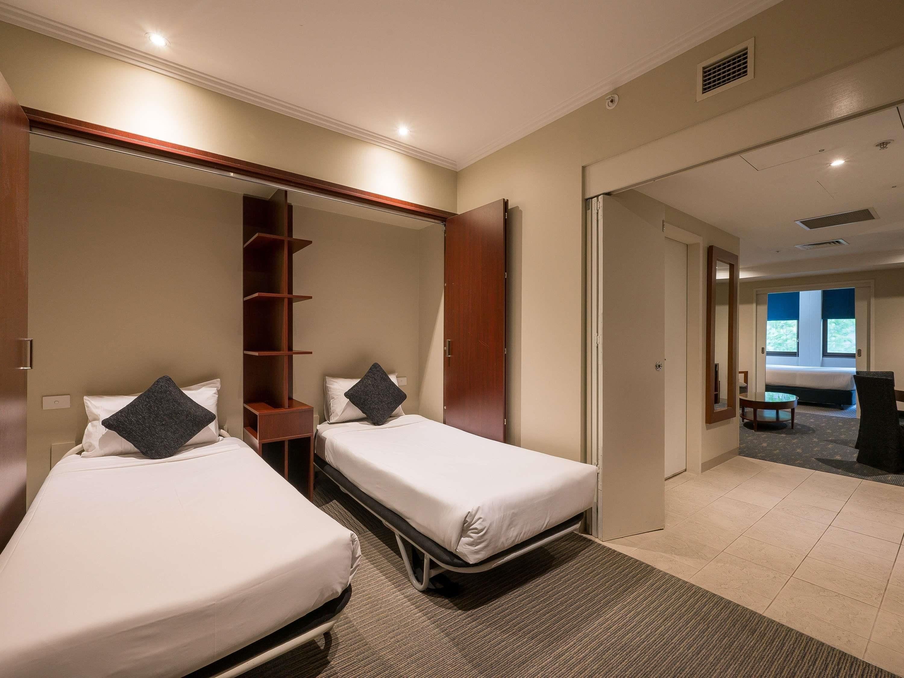 A hotel room with two beds, a wooden shelf unit, and a view of the outdoors through a window.