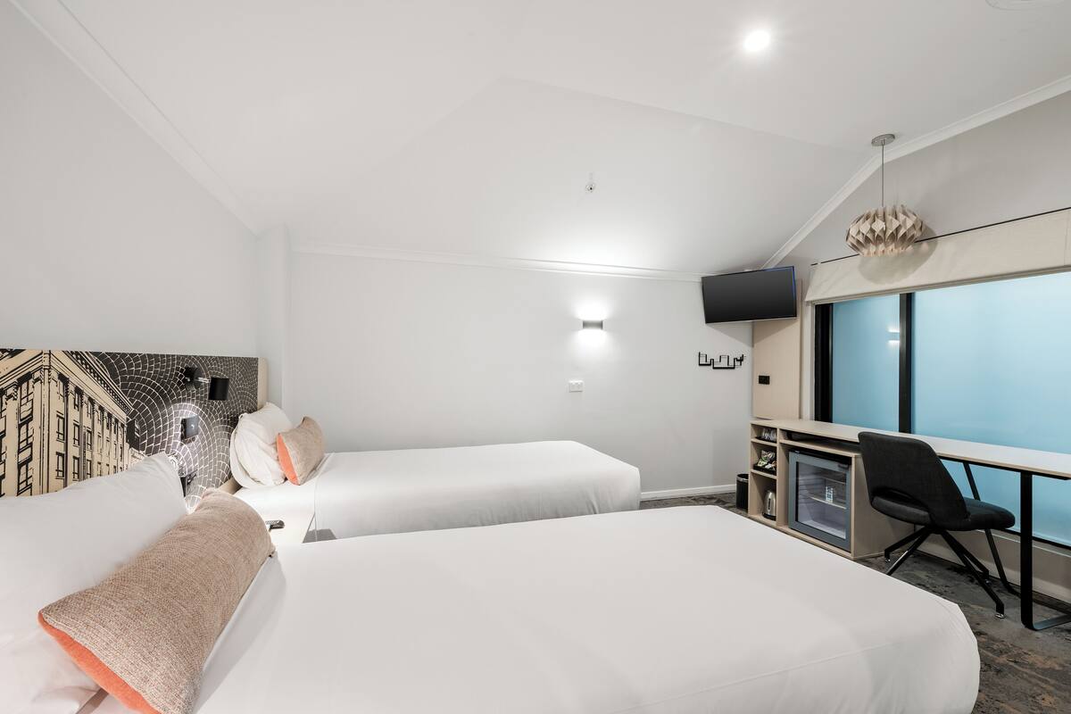 Quarto de hotel com duas camas, uma mesa com cadeira, televisão e uma janela grande.