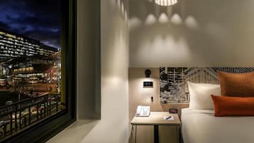 Habitación de hotel con cama, mesita de noche con teléfono y reloj, lámpara de pared e vista panorámica de la ciudad a través de la ventana.