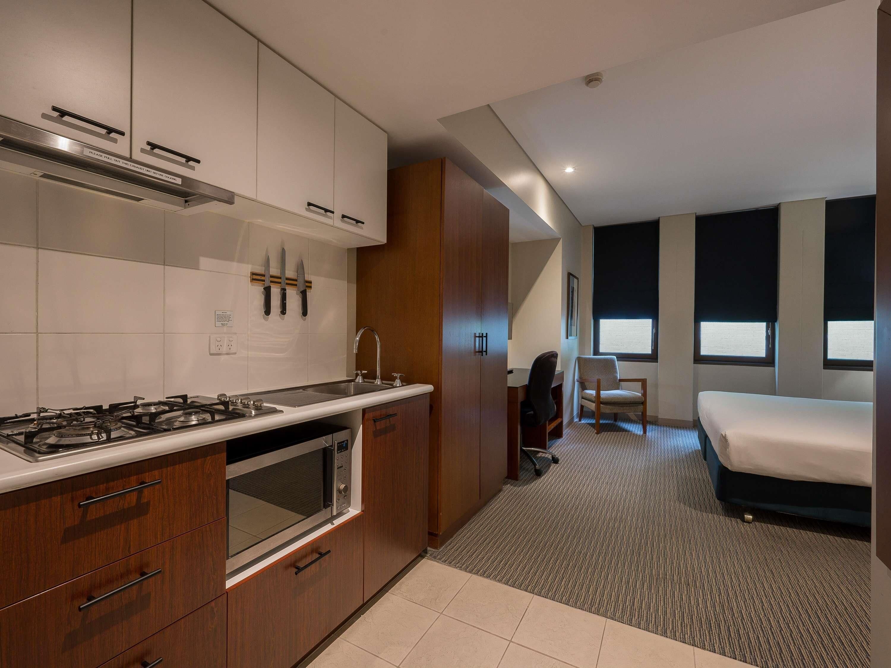 Cocina compacta con estufa, pileta y alacenas, un pequeño espacio de trabajo con una silla, y una cama en una habitación con ventanas amplias.