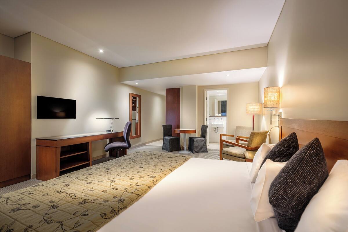 Quarto de hotel com cama, escrivaninha, cadeira, televisão e luminárias.